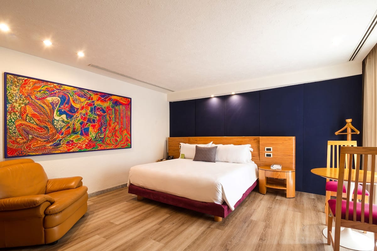 Ein Hotelzimmer mit einem großen Bett, einem hölzernen Kopfteil, einem farbenfrohen abstrakten Gemälde an der Wand, einem braunen Ledersessel und einem kleinen Holztisch mit Stuhl.