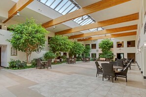 A spacious indoor area with high ceilings, wooden beams, and large windows allowing natural light. There are multiple seating areas with wicker furniture and potted plants. - Best Western Pecos Inn (Artesia)