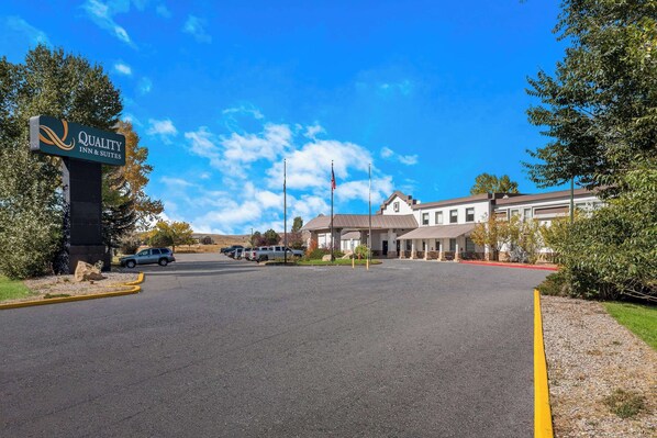 A hotel entrance with a sign reading "Quality Inn & Suites," a parking lot, and a building with a flagpole.