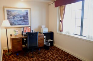 A room with a desk, chair, lamp, framed painting, printer, and a window with curtains.