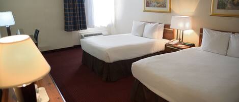 Habitación de hotel con dos camas, un escritorio con lámpara, una ventana con cortinas y dos cuadros en la pared.