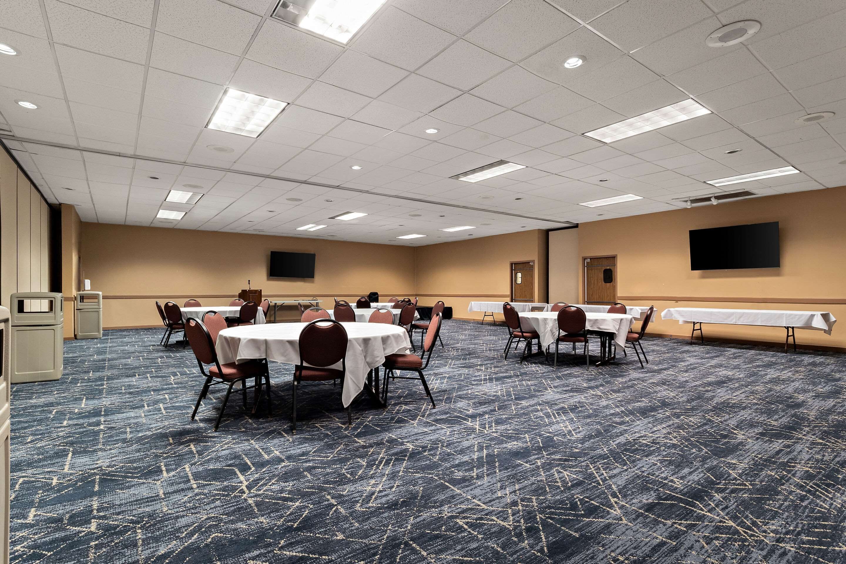 A conference room with tables and chairs, a flat-screen TV mounted on the wall, and a carpeted floor.