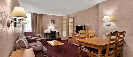 A hotel room with a living area featuring a sofa set, a wooden dining table with chairs, a TV, and wall-mounted lamps.