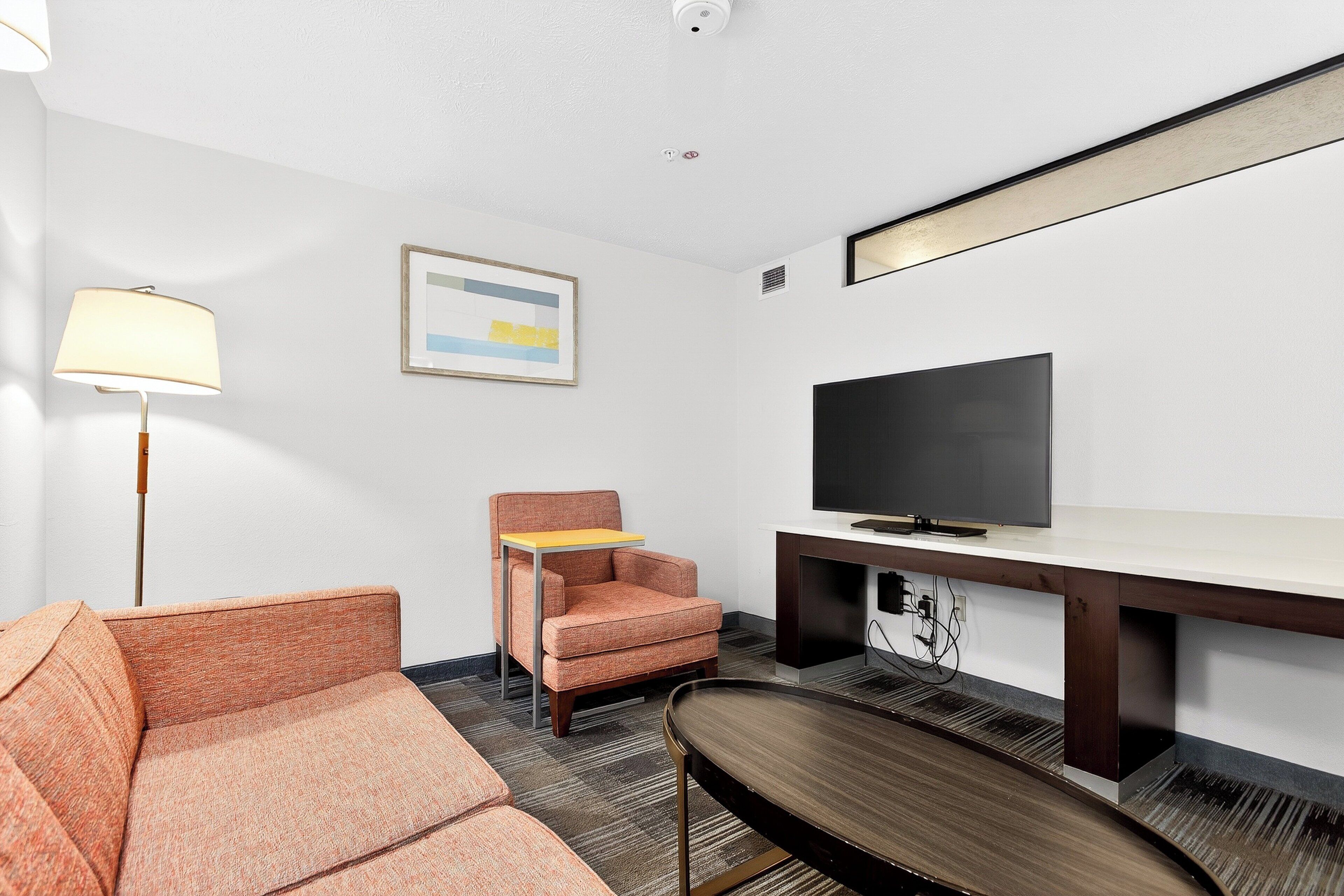 A hotel room with a sofa, armchair, TV, desk, lamp, and artwork on the wall.