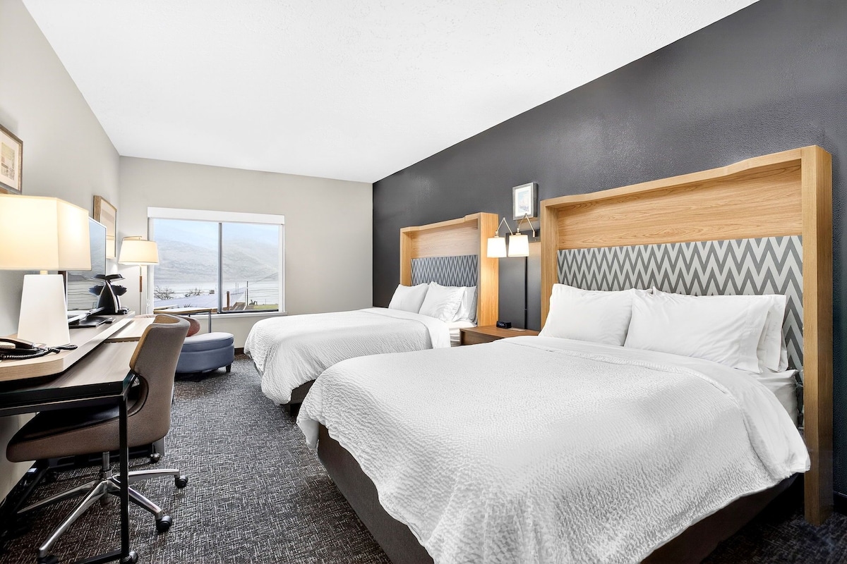 A hotel room with two beds, a desk, a chair, a lamp, and a view of the mountains.