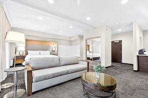 A hotel room with a sofa, a round glass coffee table, a lamp, a mirror, and a desk.