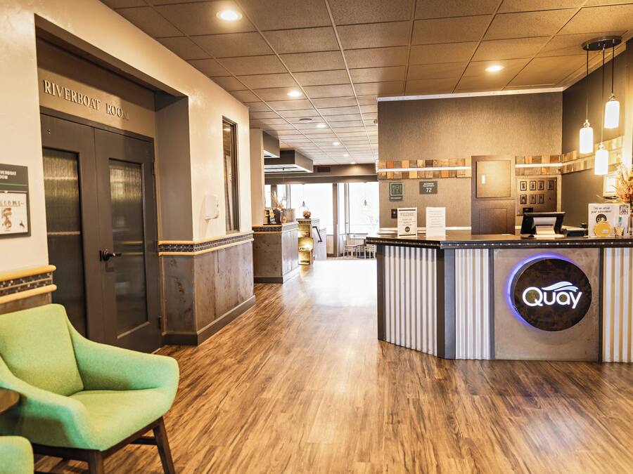 A reception area with a Riverboat Room sign, a reception desk with a 'Quay' logo, a green chair, and a wooden floor.