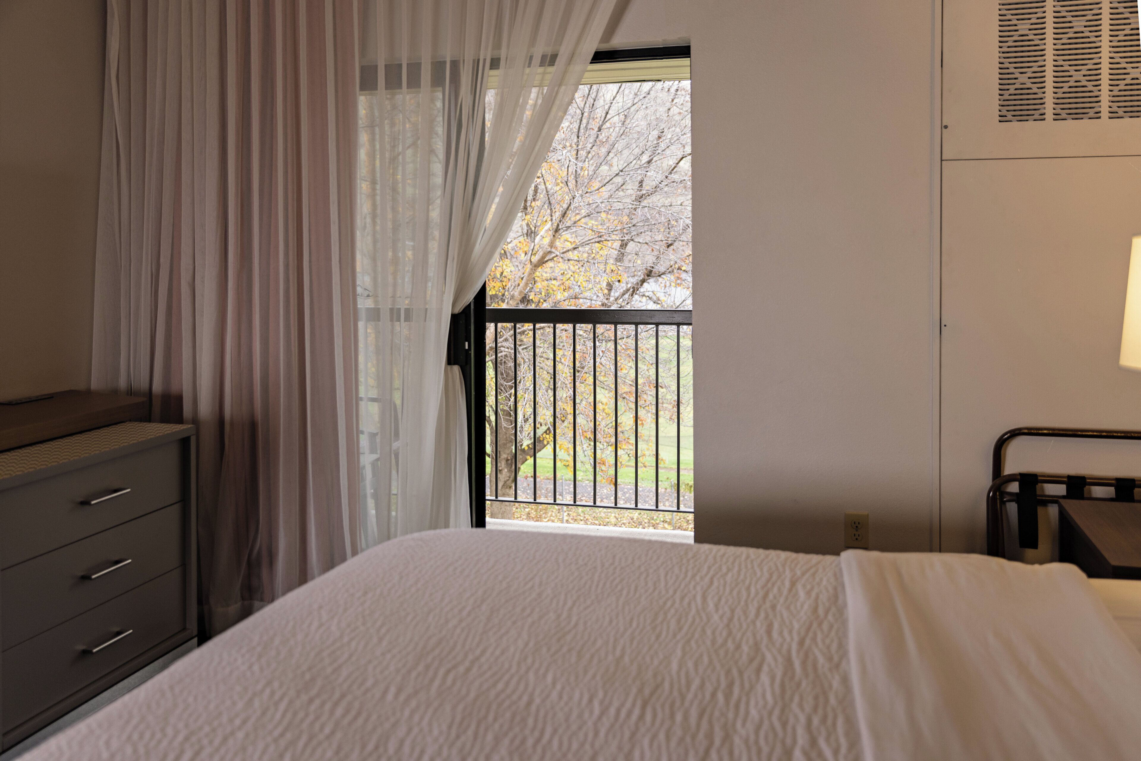 A bedroom with a bed, a dresser, a window with a view of trees, and a balcony railing.