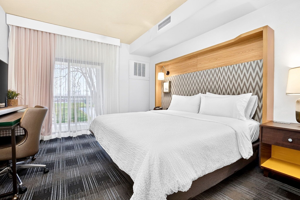 A hotel room with a large bed, a desk with a chair, a TV, and a window with curtains.
