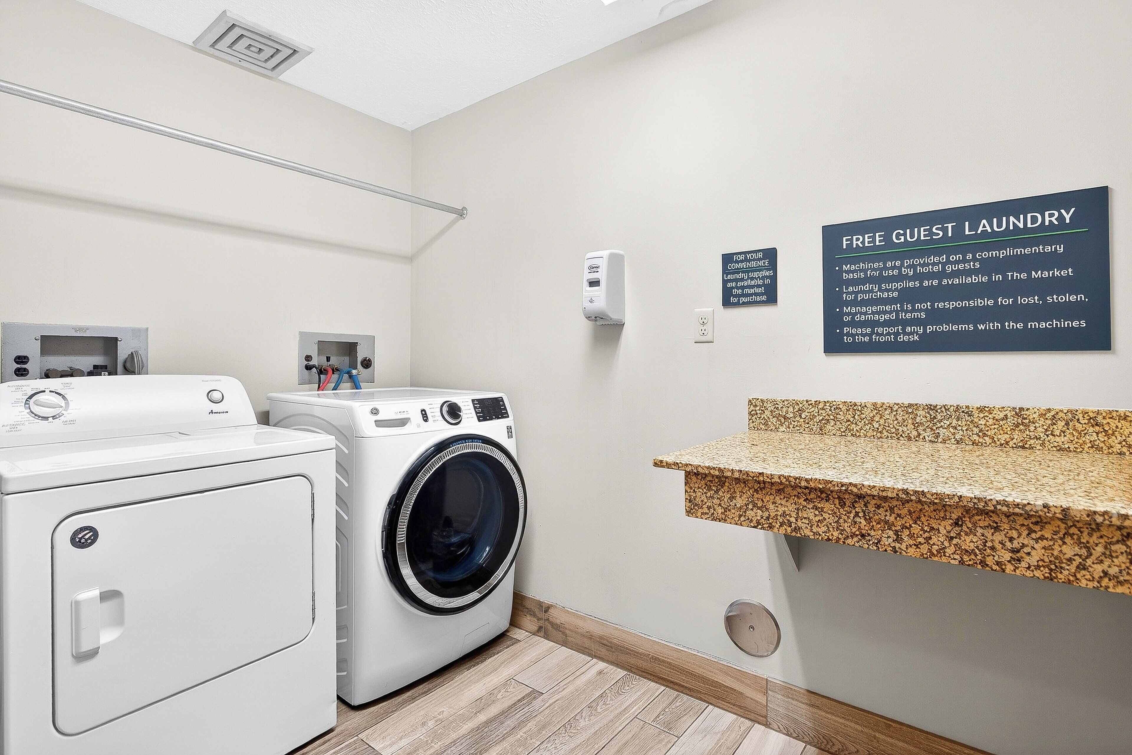 A hotel laundry area with two washing machines, a drying rack, a soap dispenser, and a granite countertop.