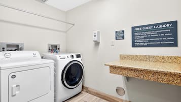 A hotel laundry area with two washing machines, a drying rack, a soap dispenser, and a granite countertop.