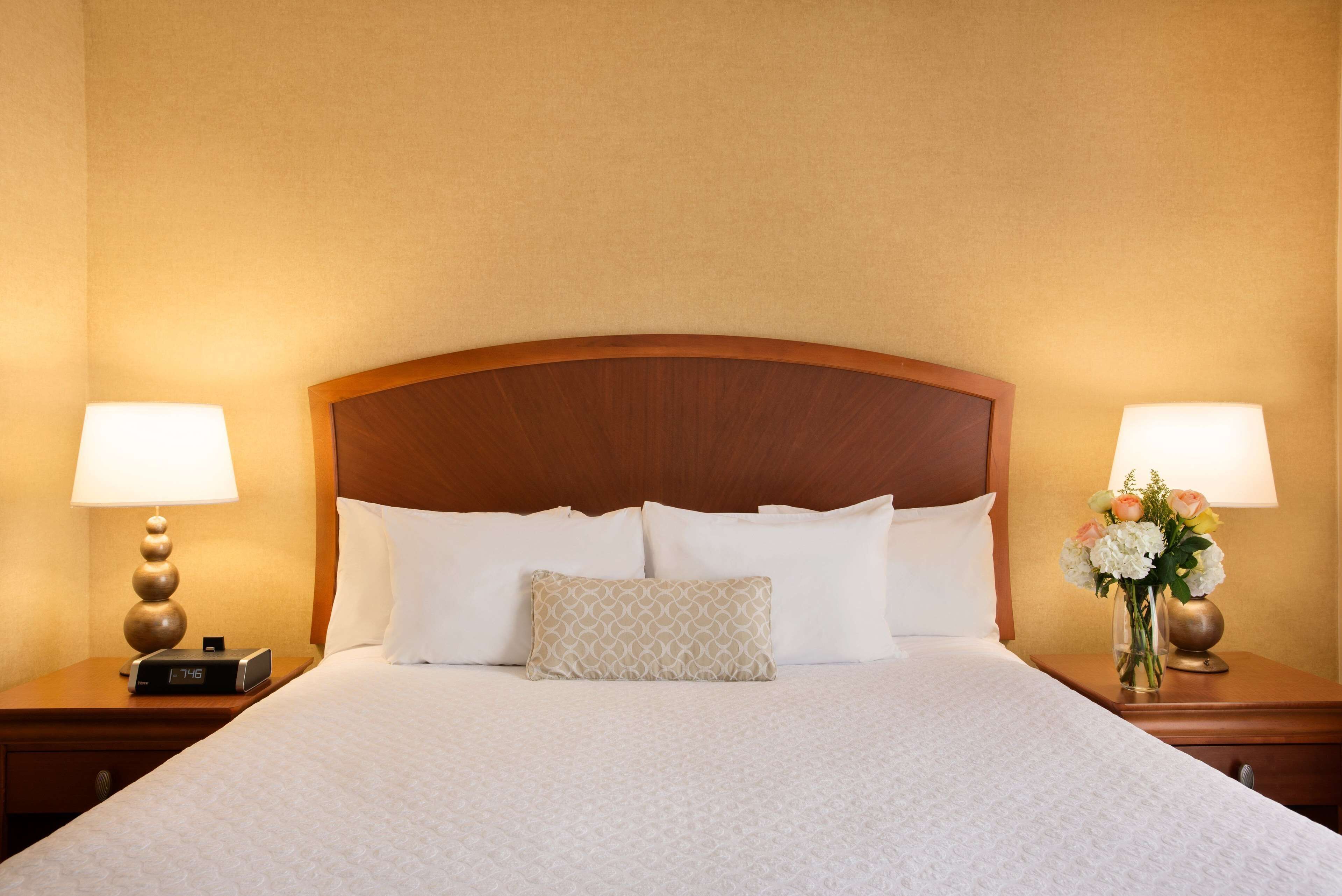 Una habitación de hotel con una cama grande, dos mesitas de noche con lámparas, un reloj y un jarrón con flores.
