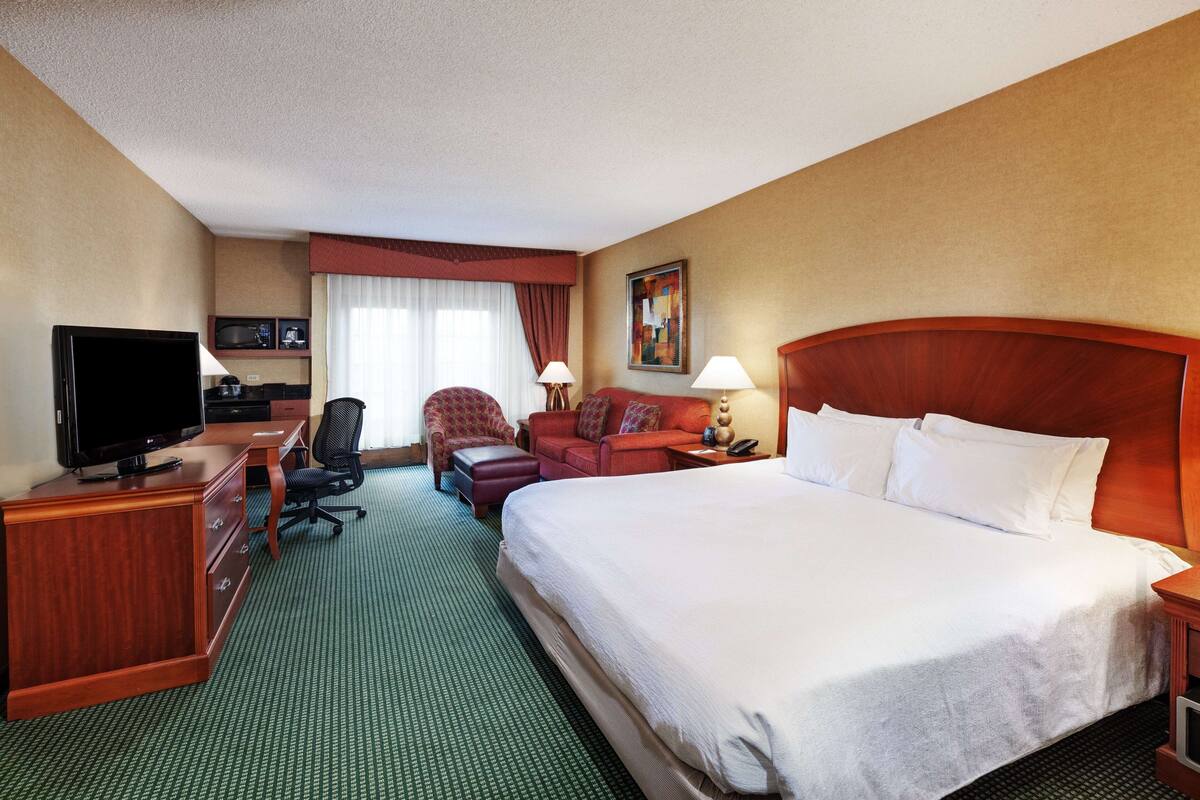 A hotel room with a large bed, a desk with a TV, a chair, a sofa, and a painting on the wall.