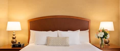 Una cama bien hecha con sábanas blancas y cabecera de madera, flanqueada por dos mesitas de noche con lámparas y un florero.