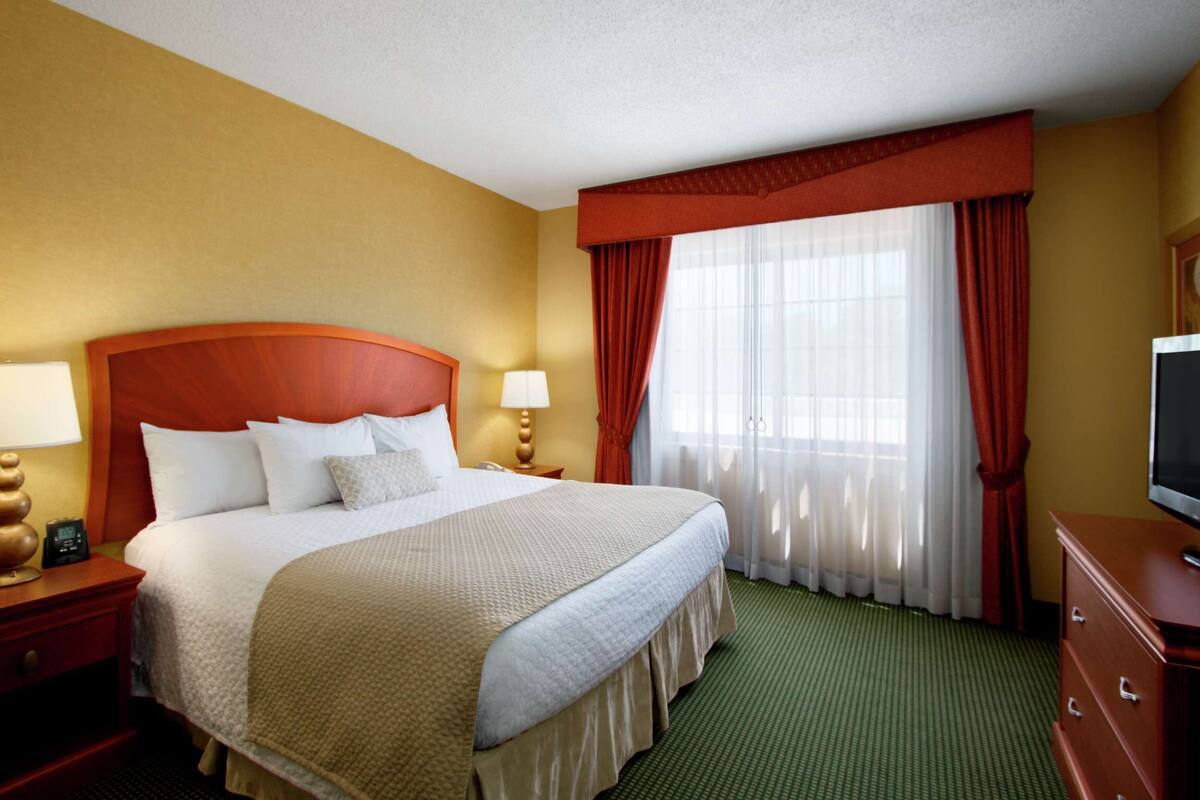 Une chambre d’hôtel avec un grand lit, deux tables de chevet avec des lampes, un téléviseur posé sur une commode et une fenêtre avec des rideaux rouges.