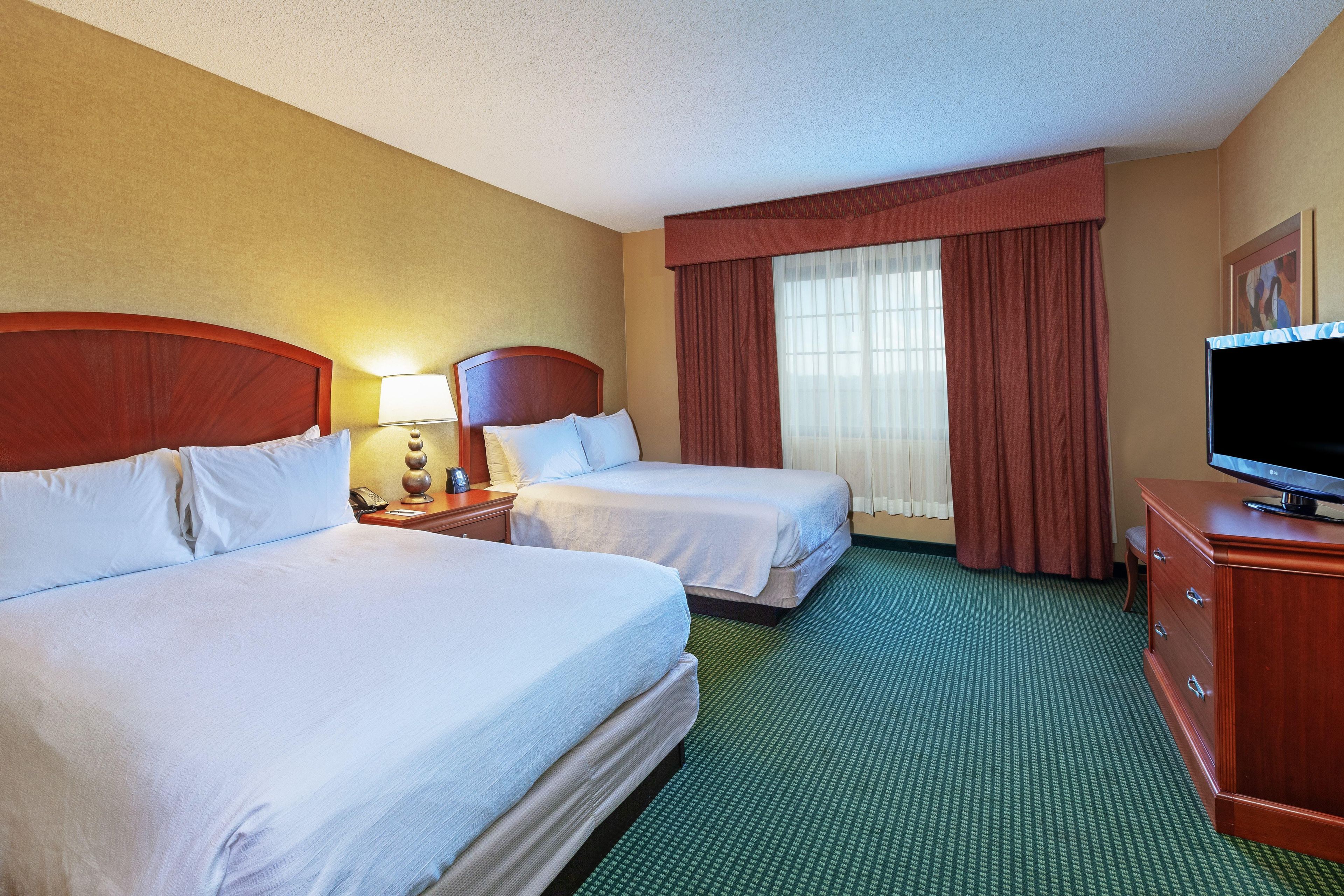 Ein Hotelzimmer mit zwei Betten, einem Fernseher, einem Schreibtisch mit Lampe und einem Fenster mit Vorhängen.