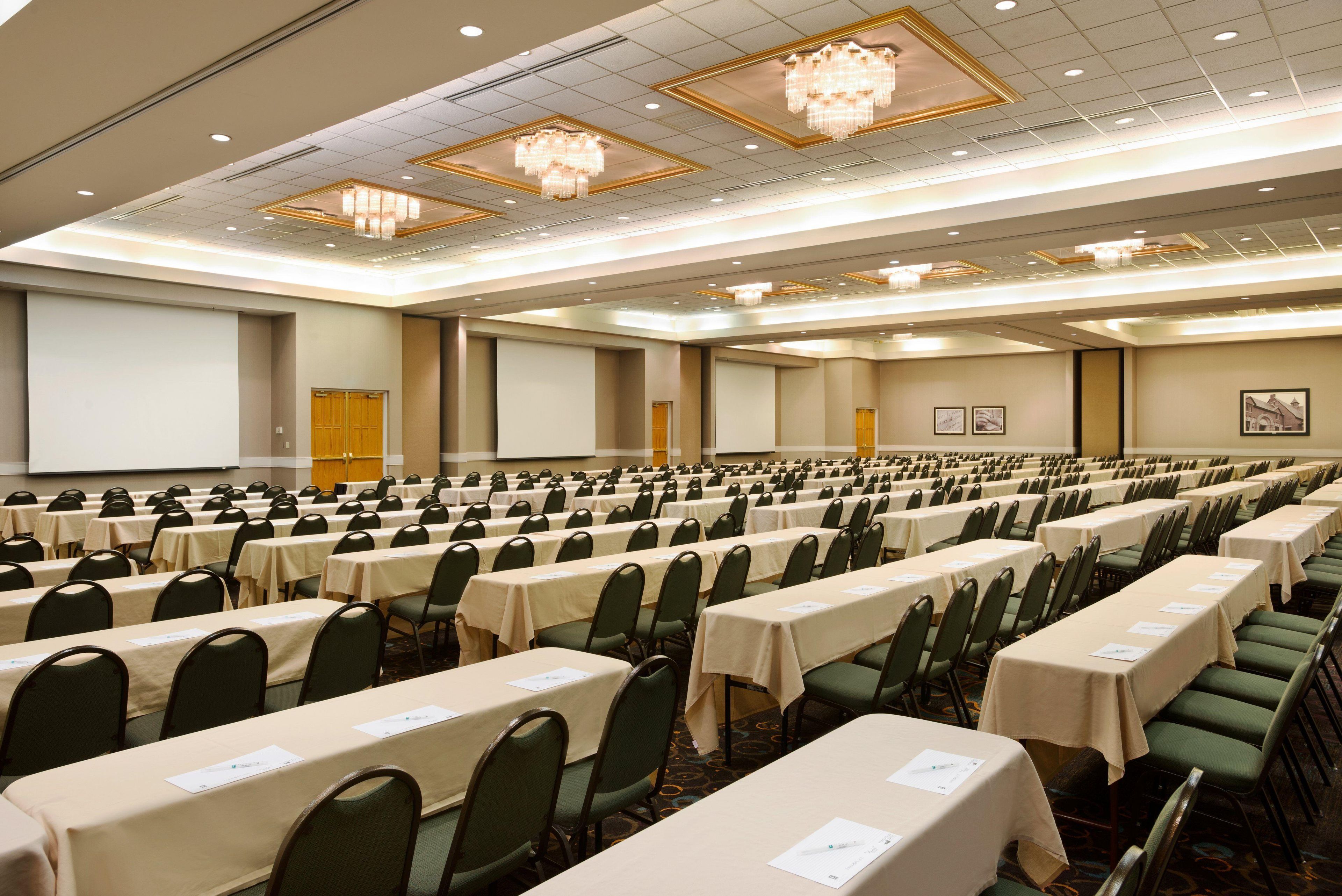 A large conference room with rows of tables and chairs, a stage with a projector screen, and a chandelier.