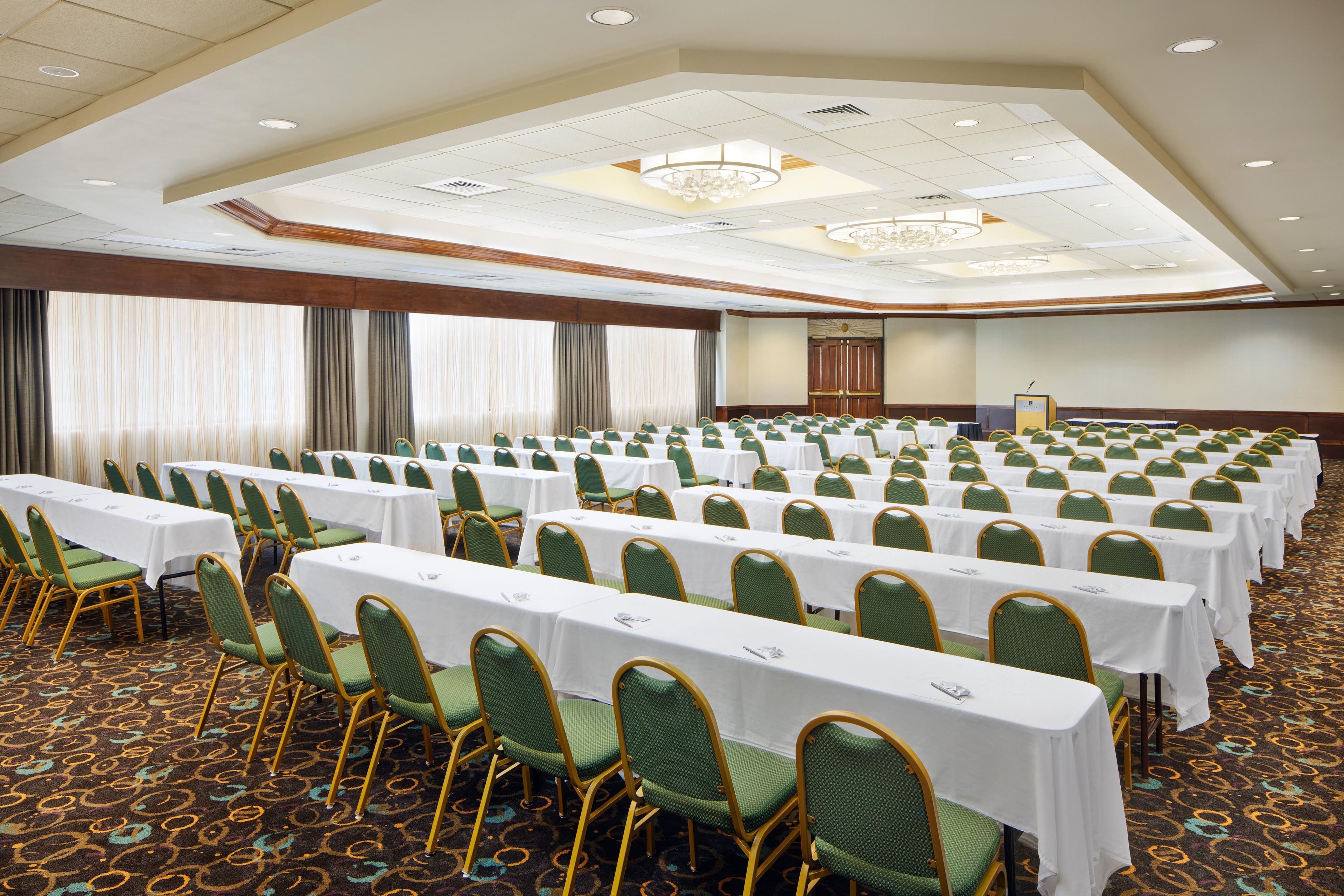 A conference room with rows of tables and chairs, a podium, and a large window with curtains.