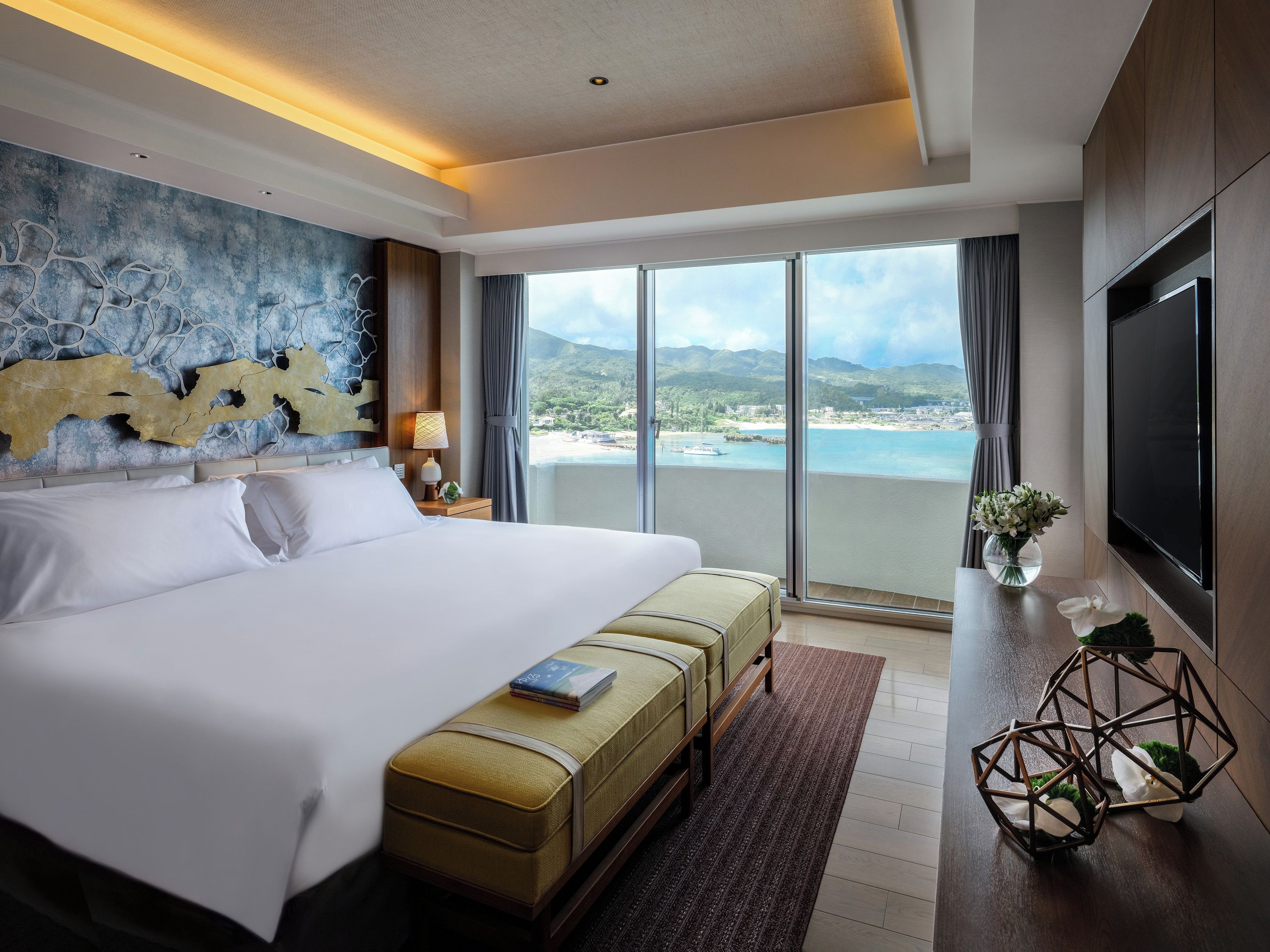 Habitación de hotel con una cama grande, vista a la playa, un televisor de pantalla plana y una pequeña mesa con un florero.