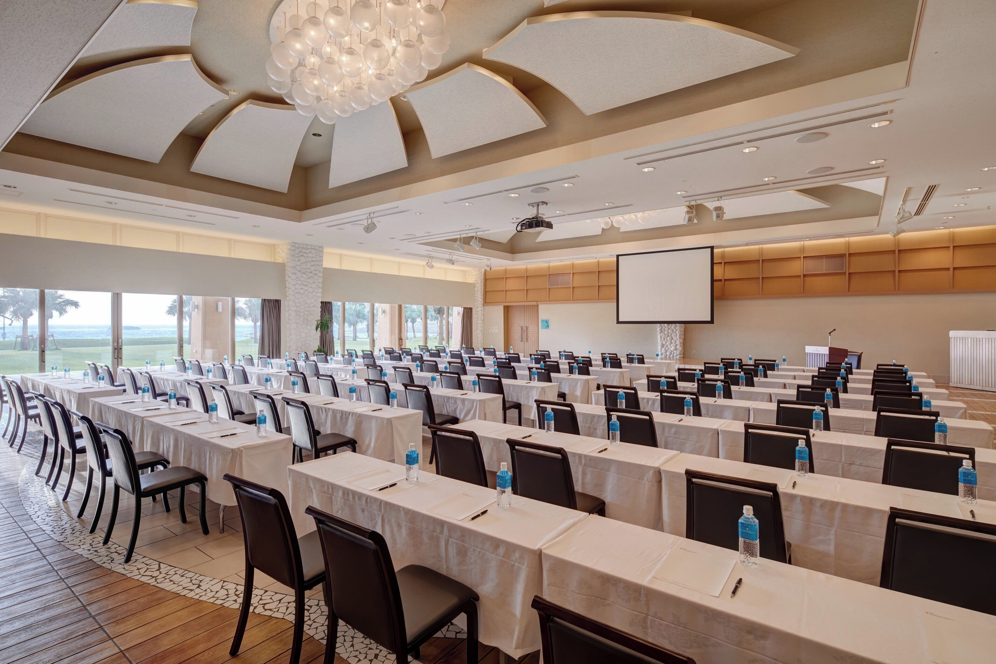 A conference room with multiple tables and chairs, a projector screen, and a large window with a view of the outdoors.