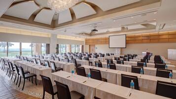 A conference room with multiple tables and chairs, a projector screen, and a large window with a view of the outdoors.