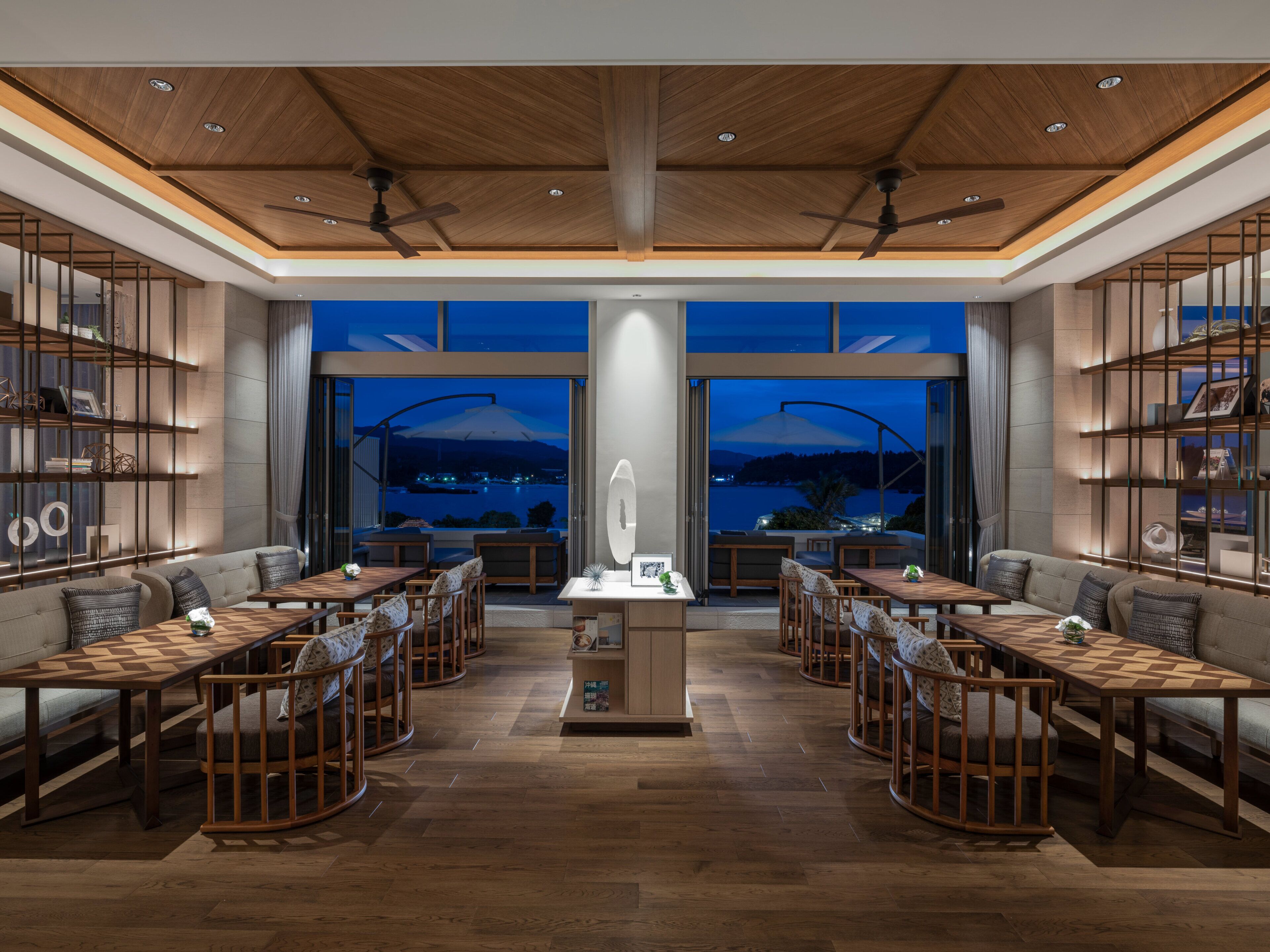 A modern hotel lobby with a central reception desk, seating areas with tables and chairs, and large windows offering a view of the cityscape at night.