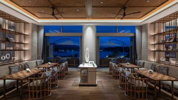 A modern hotel lobby with a central reception desk, seating areas with tables and chairs, and large windows offering a view of the cityscape at night.