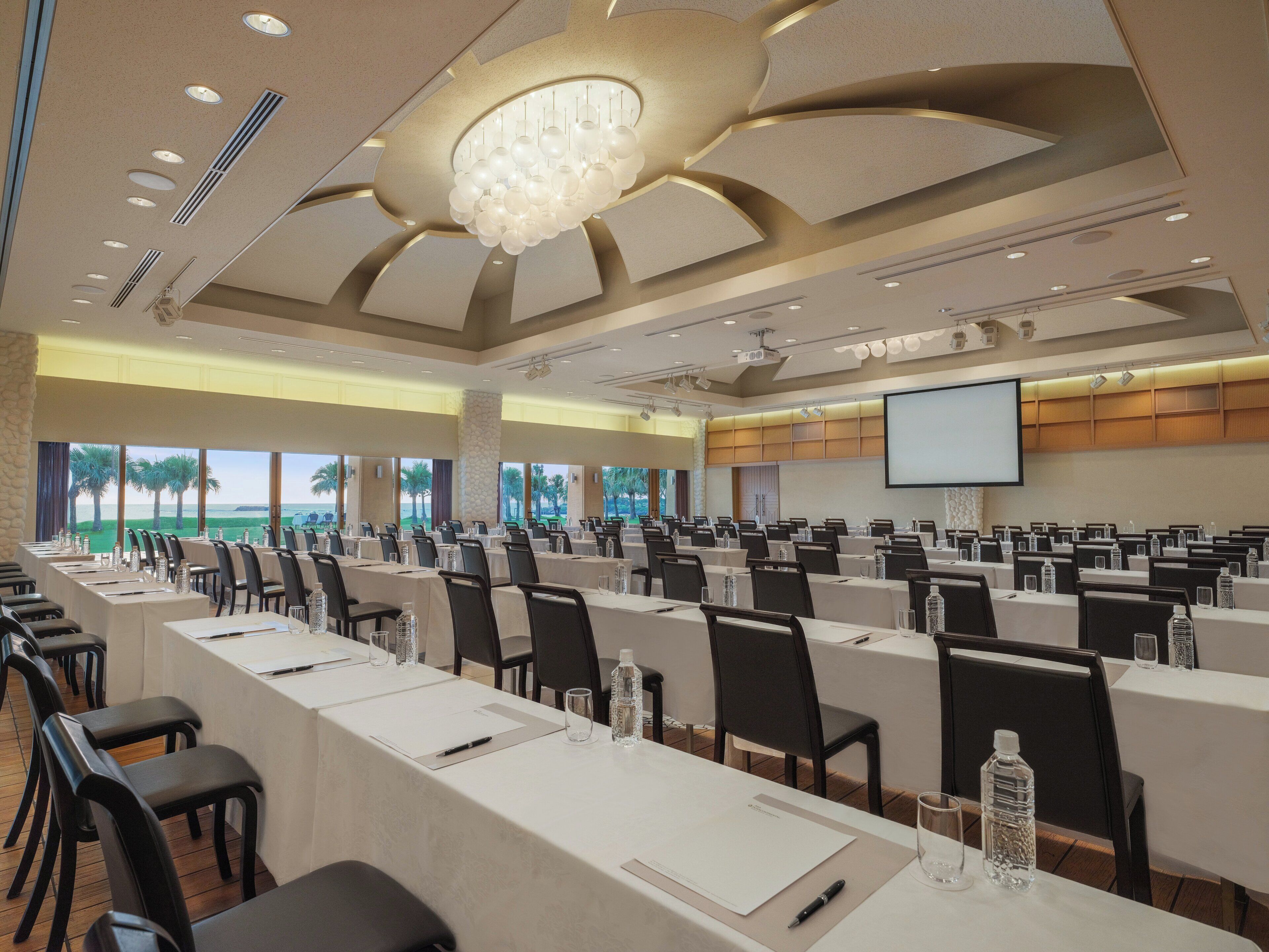 A conference room with multiple tables, chairs, a large screen, and a view of the outdoors.