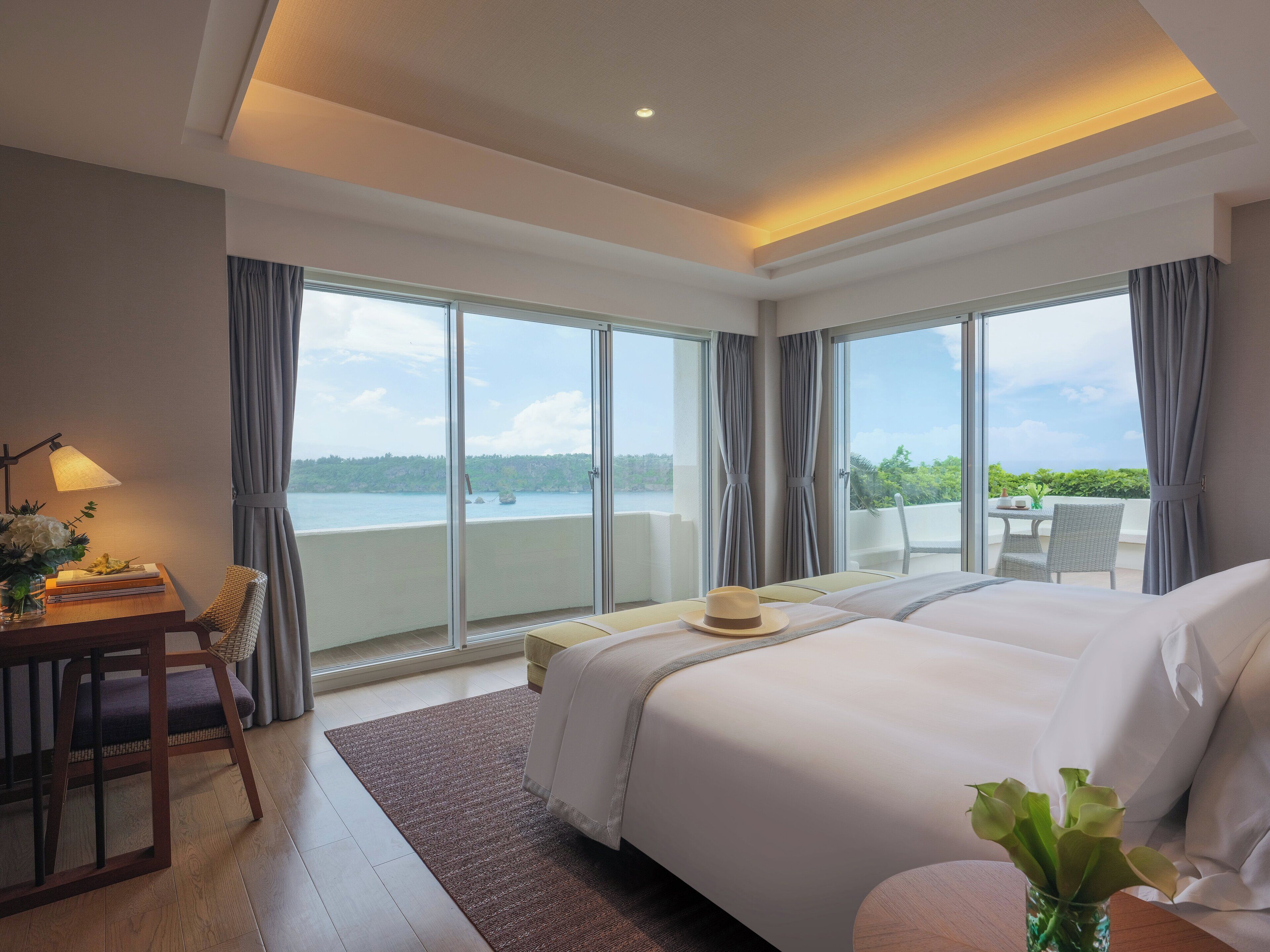 A hotel room with a large bed, a desk with a lamp, and a view of the sea through large windows.