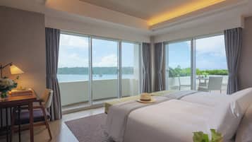 A hotel room with a large bed, a desk with a lamp, and a view of the sea through large windows.