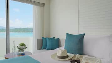 Una habitación de hotel con una cama, una ventana con vista al mar, una mesa con un sombrero, gafas y un florero con flores.
