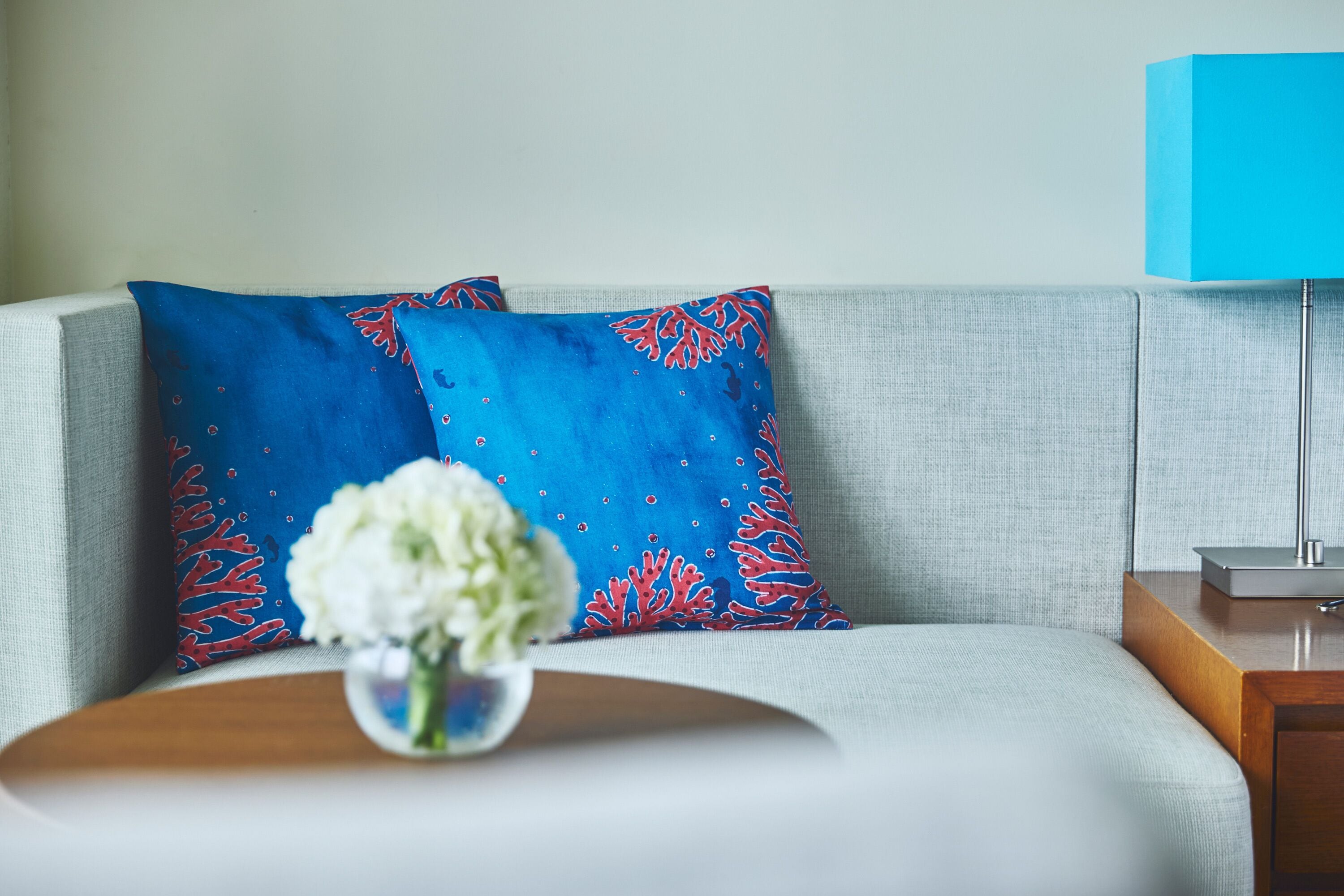 A modern living room with a light grey sofa, a blue patterned pillow, a small white flower vase, and a blue lamp on a wooden side table.