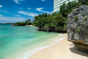 Una playa con agua turquesa cristalina, una orilla arenosa y un edificio blanco grande con balcones con vistas al mar.
