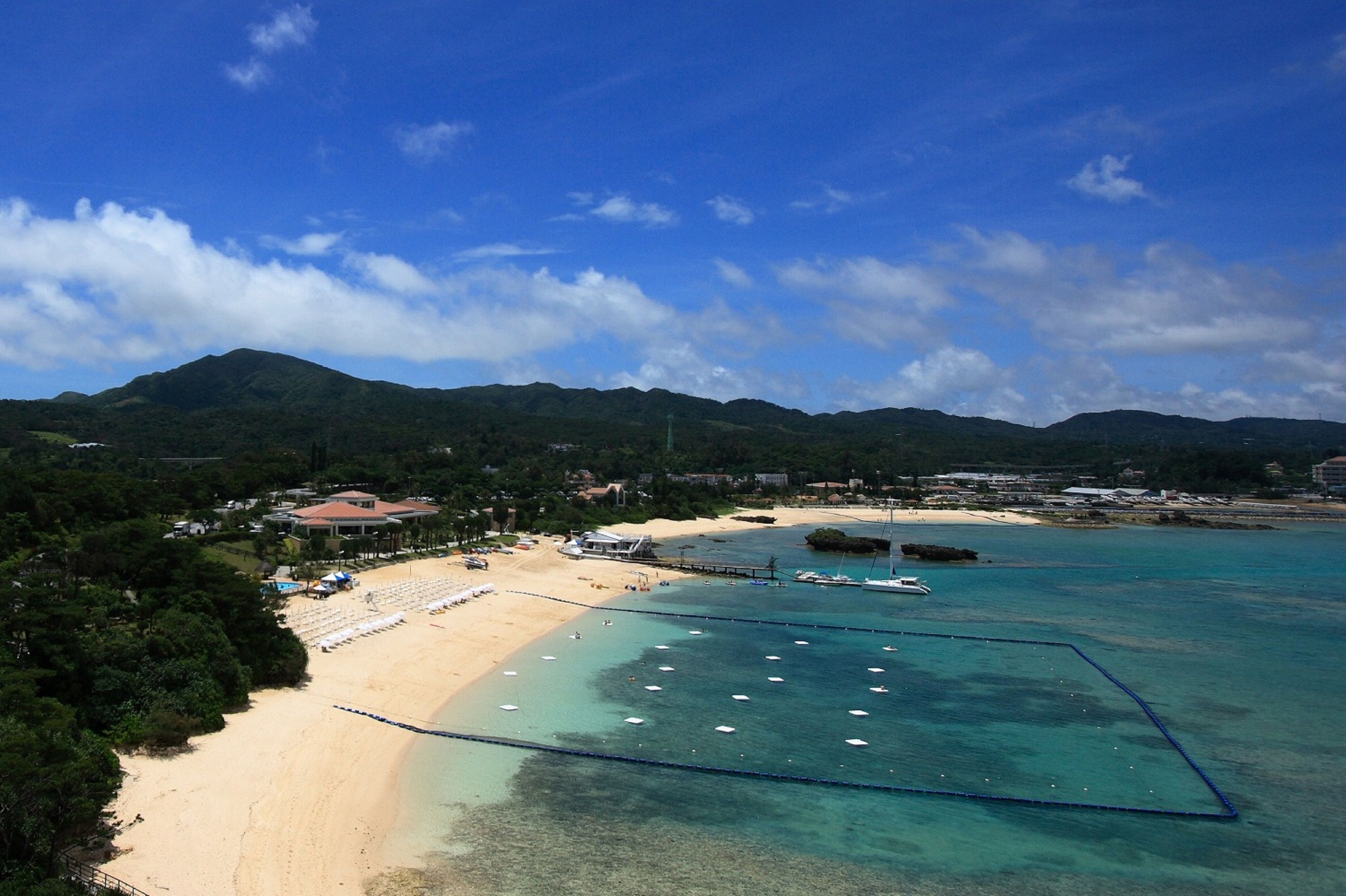 Une plage aux eaux turquoise et limpides, une jetée et un paysage montagneux en arrière-plan.