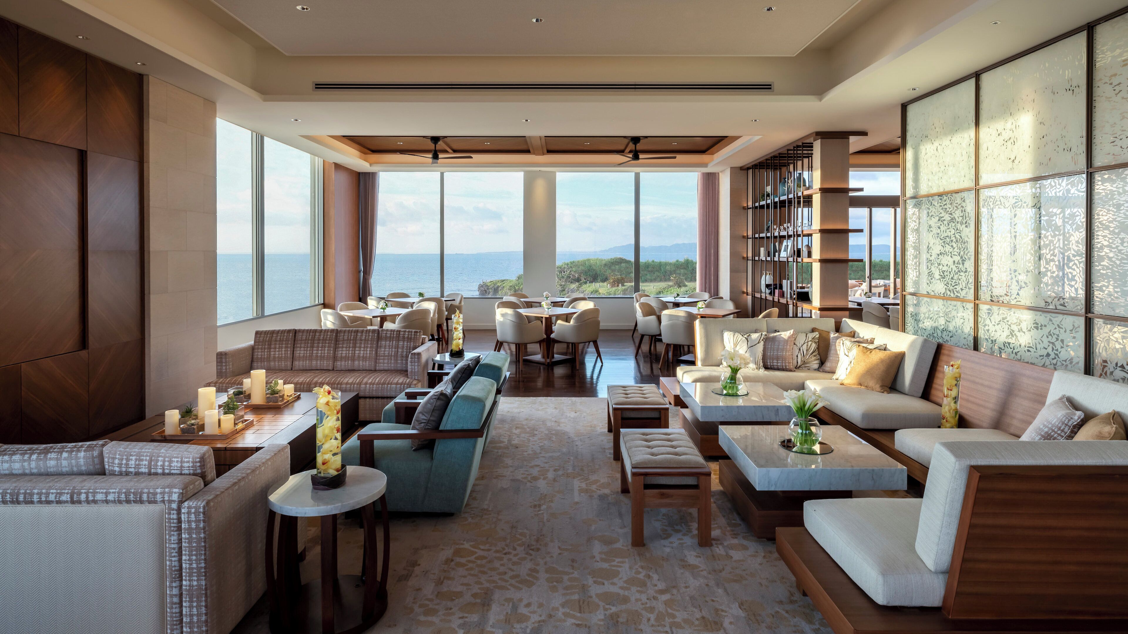A spacious lounge area with multiple seating options, large windows offering a view of the sea, and a ceiling fan.