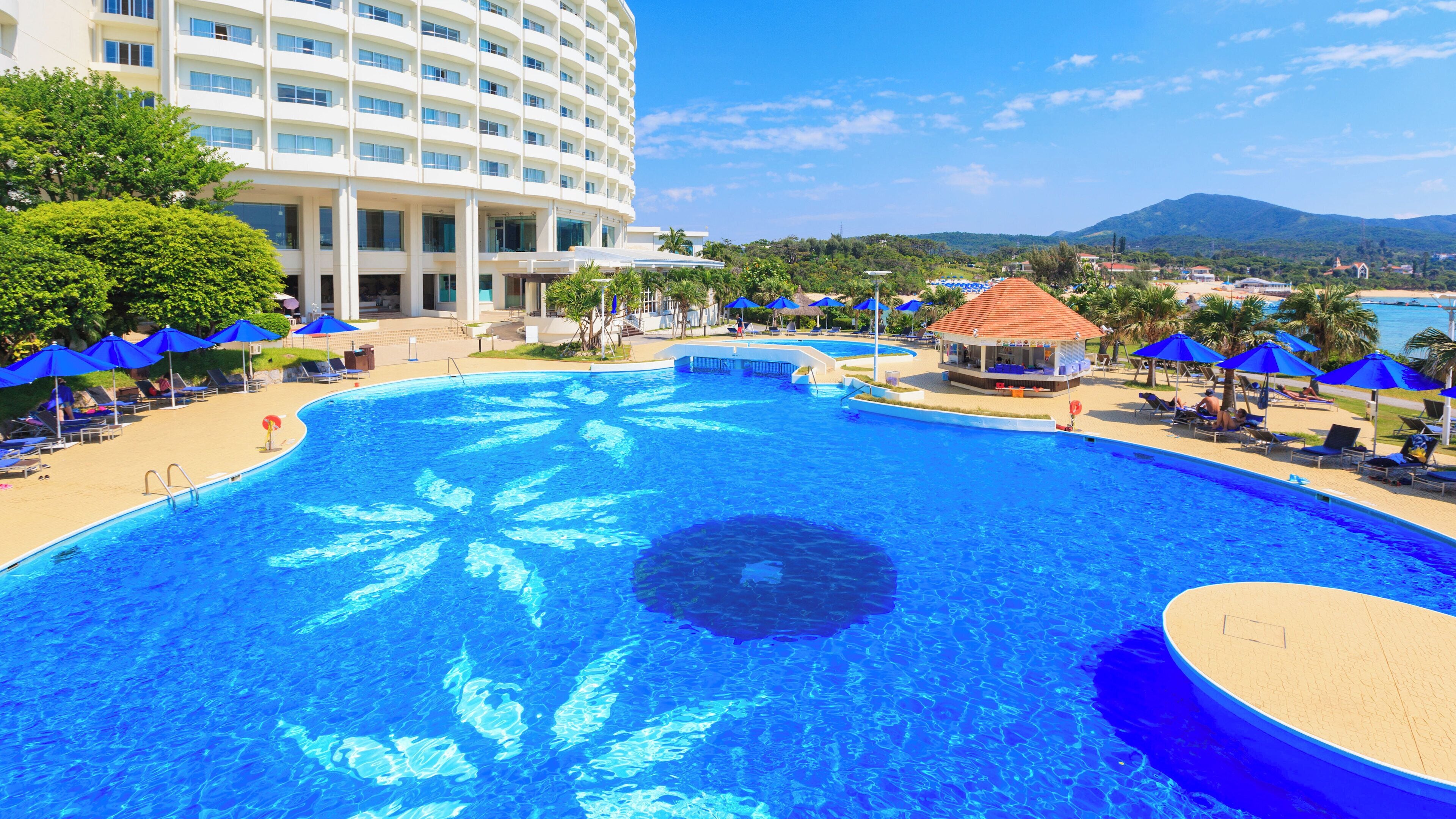 A large swimming pool with a tropical design, surrounded by lounge chairs and umbrellas, with a hotel building in the background.
