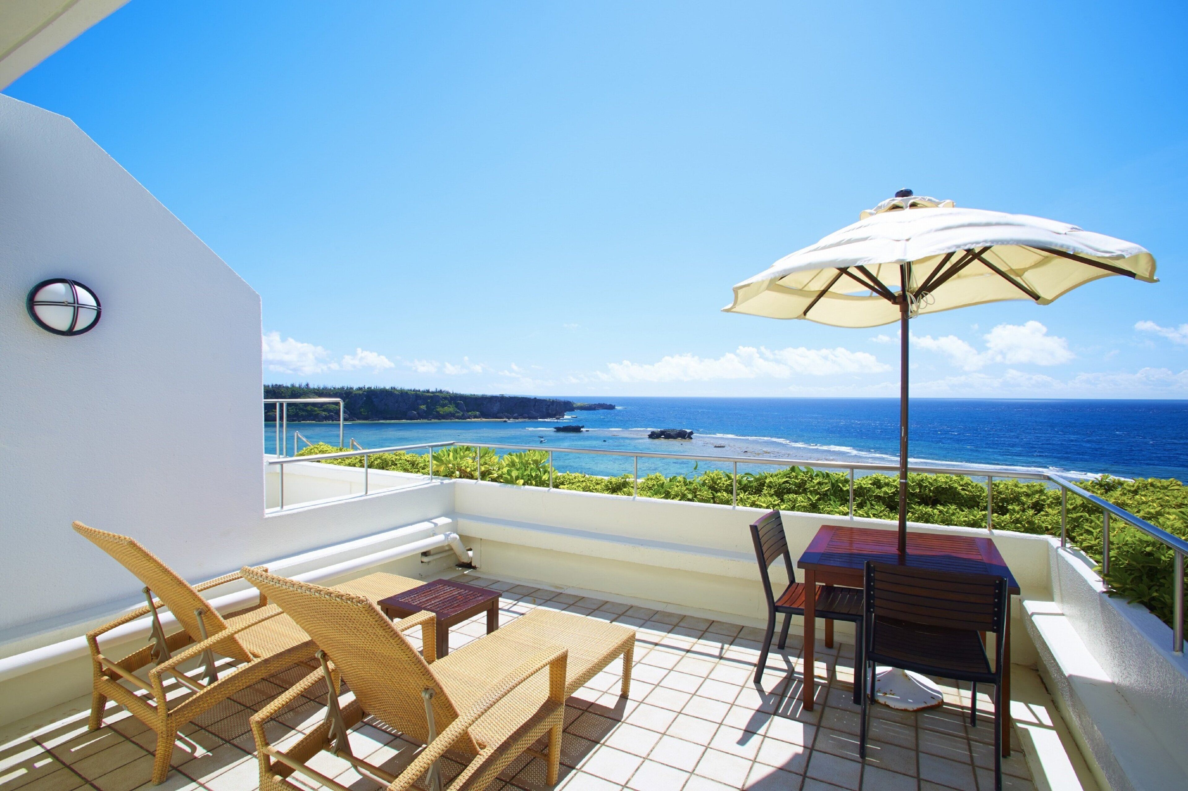 A balcony with wicker chairs, a table, and an umbrella, overlooking a coastal view with the ocean and a distant island.