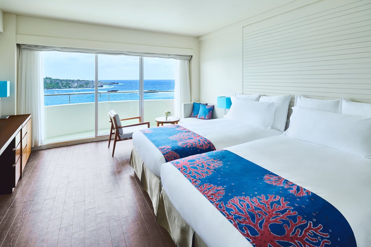 A hotel room with two beds, a balcony with a view of the sea, a desk, and a chair.