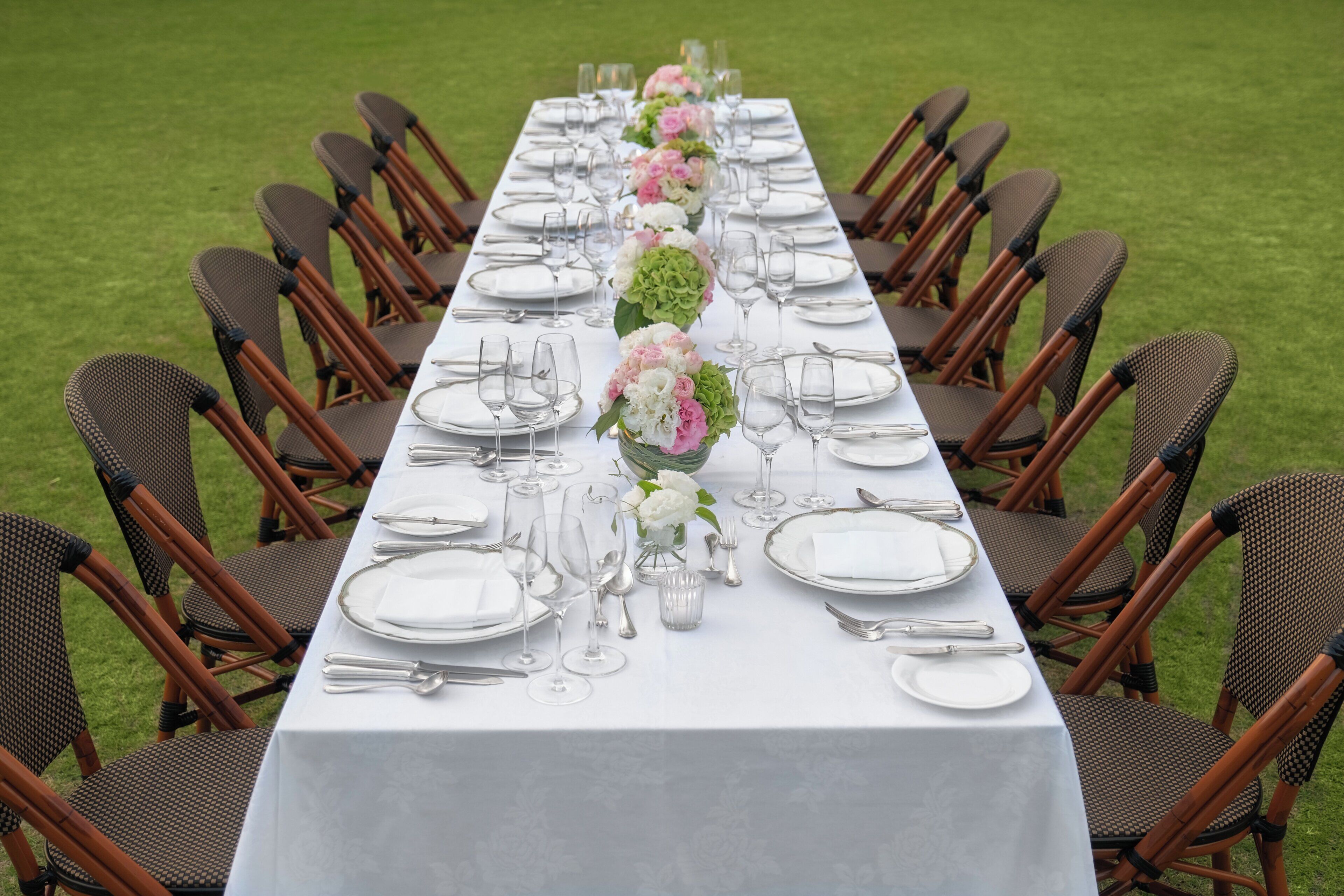 Una larga mesa de comedor dispuesta con mantel blanco, cubiertos y cristalería, flanqueada por sillas de mimbre.