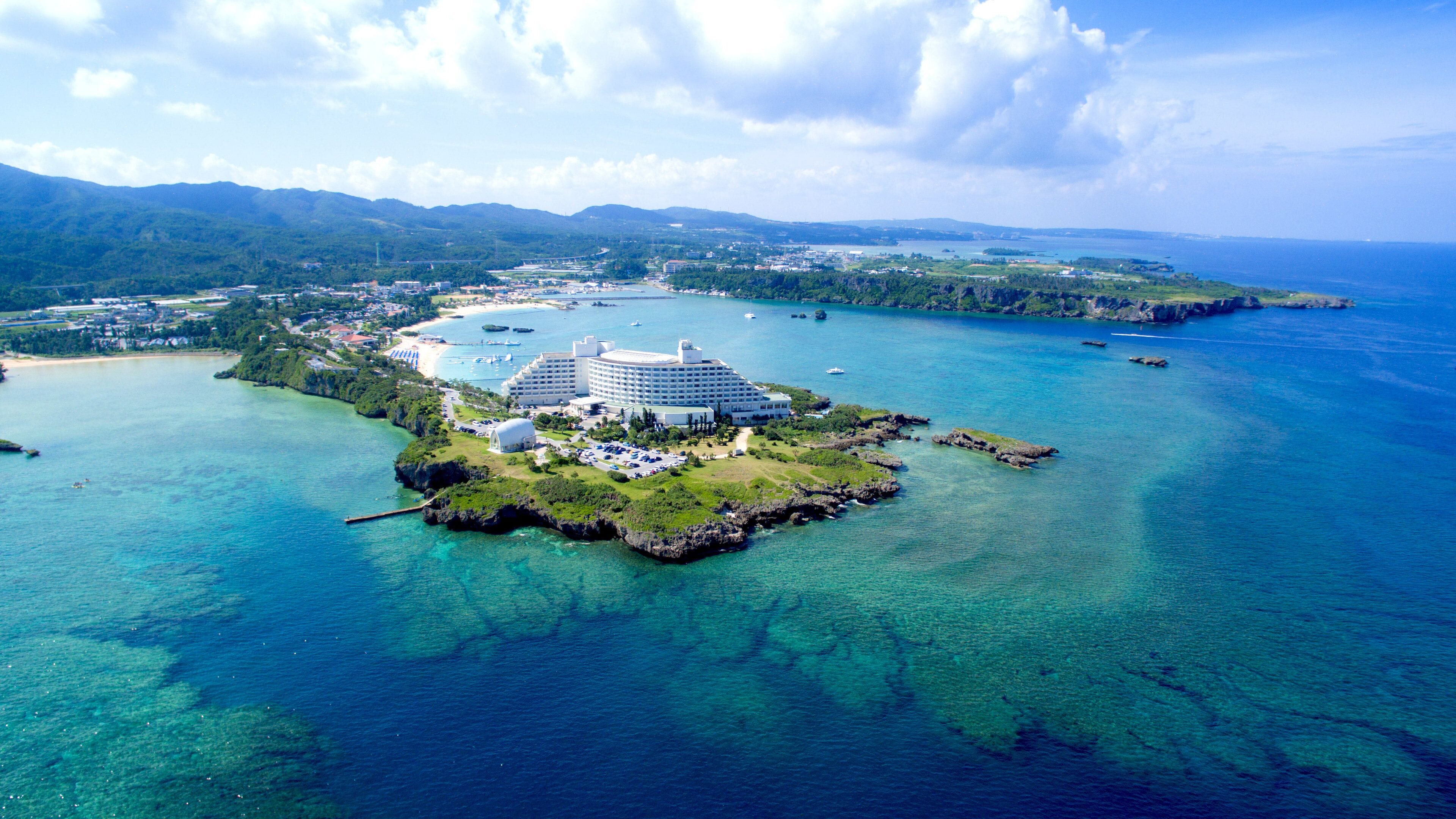 An aerial view of a resort island with a large hotel, clear blue waters, and surrounding greenery.