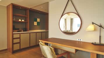 A hotel room with a wooden desk, a chair, a mirror, a lamp, and a kitchenette area.
