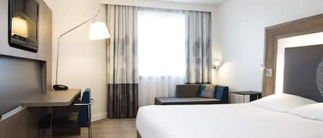 A hotel room with a bed, a desk, a chair, a lamp, and a window with curtains.