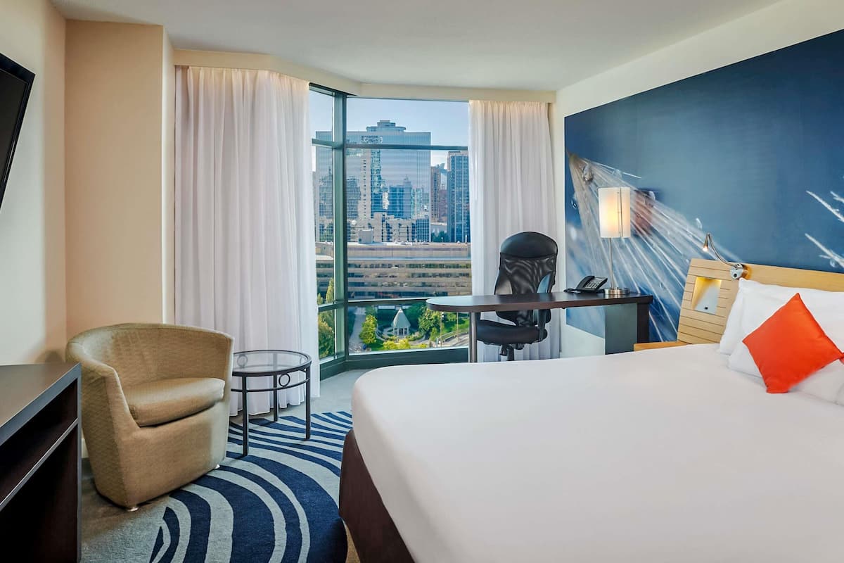 A hotel room with a bed, a desk, a chair, a TV, and a view of a cityscape through large windows.