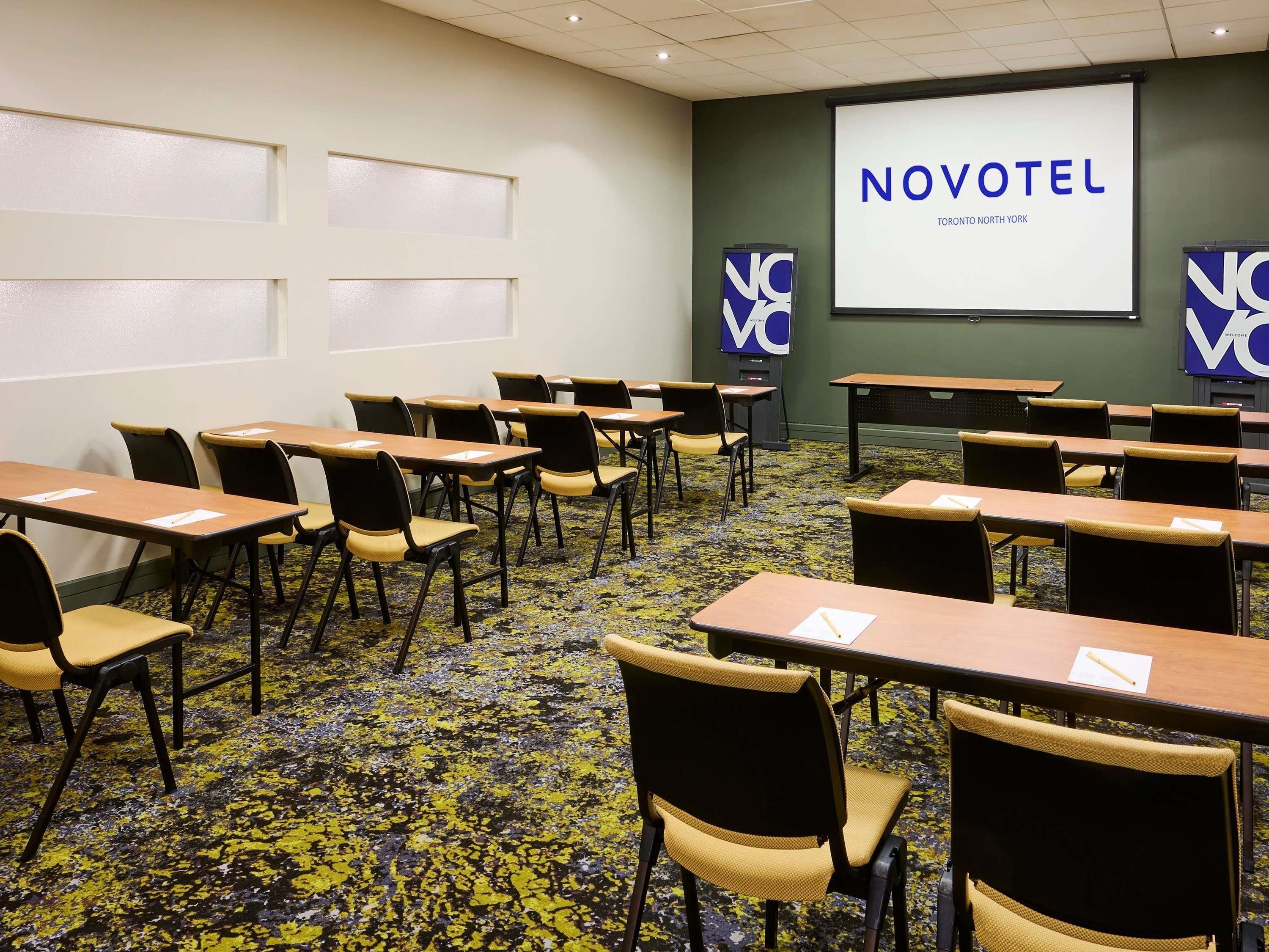 Une salle de conférence avec des rangées de tables et de chaises, un écran de projection affichant le texte « NOVOTEL TORONTO NORTH YORK », et deux panneaux « NOVOTEL » sur les murs.