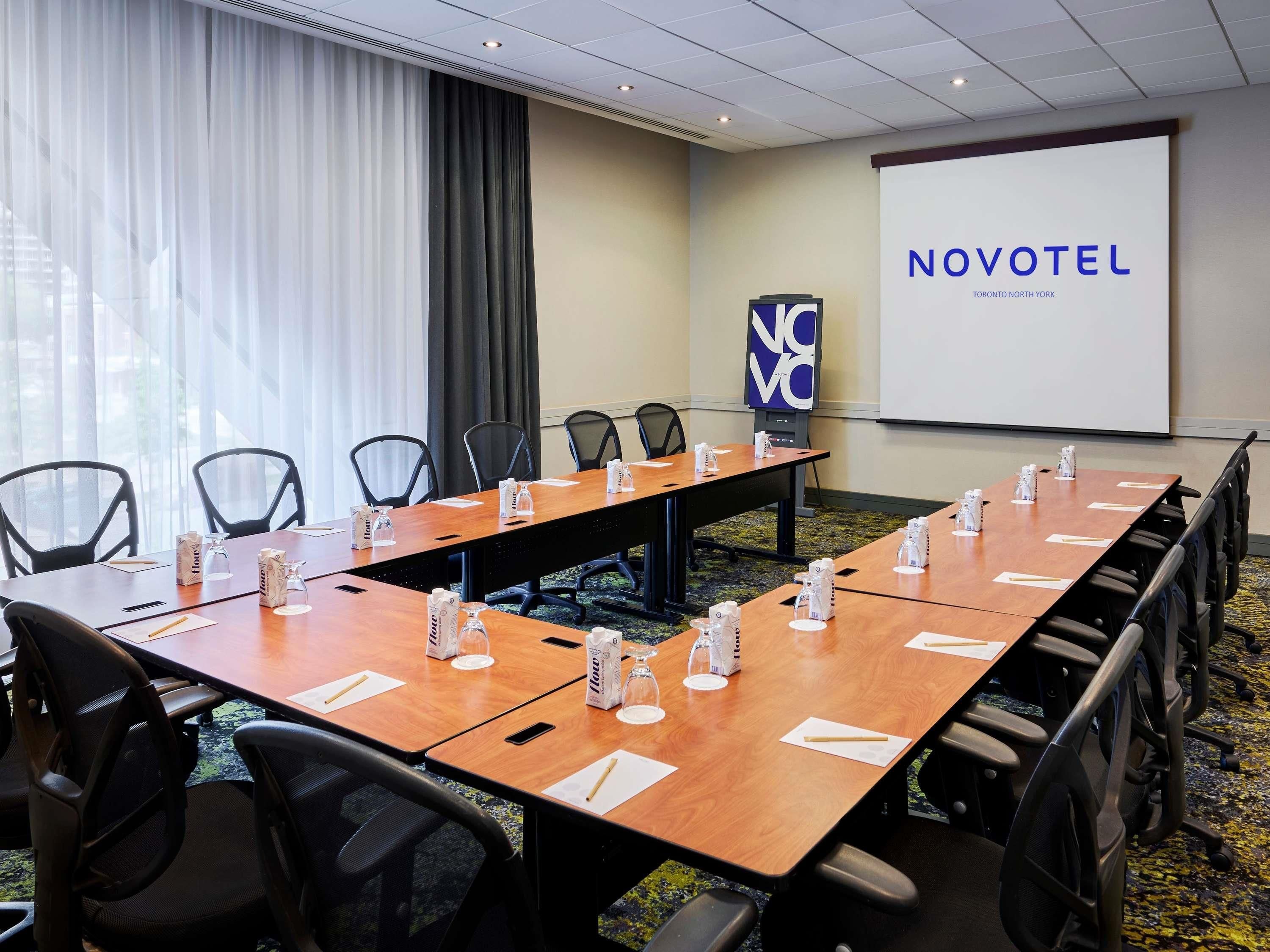 A conference room with a long table, chairs, and a projector screen displaying 'NOVOTEL Toronto North York'.