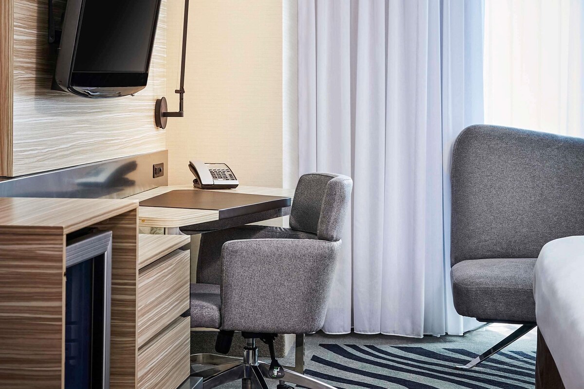 Habitación de hotel con televisor de pantalla plana montado en la pared, un escritorio con teléfono, un sillón gris y un sofá grande de color gris.