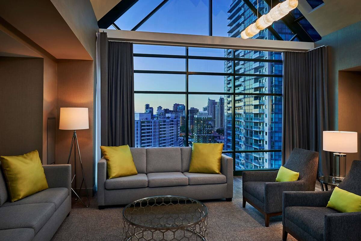 A modern hotel room with a large window offering a city view, a grey sofa with yellow cushions, a glass coffee table, and two armchairs with yellow cushions.