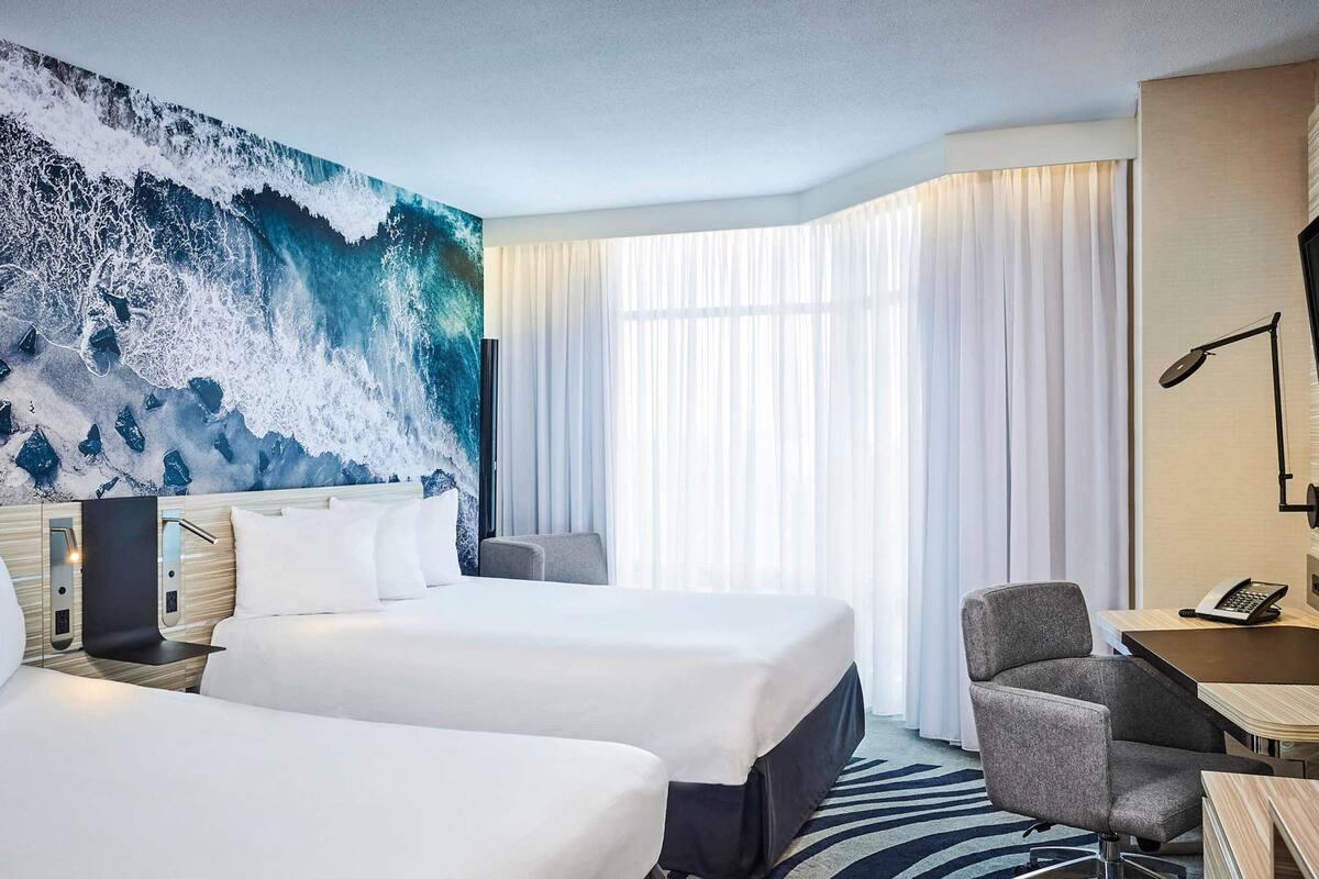 A hotel room with two beds, a large ocean mural, a desk with a lamp, and a chair.