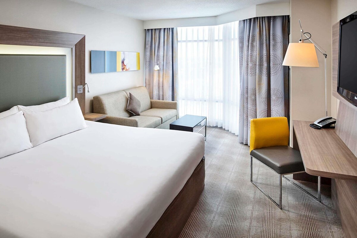 A hotel room with a large bed, a desk with a lamp, a chair, a sofa, and a TV.