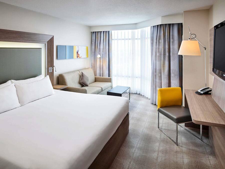 Quarto de hotel com uma cama grande, uma escrivaninha com luminária, uma cadeira, um sofá e uma televisão.